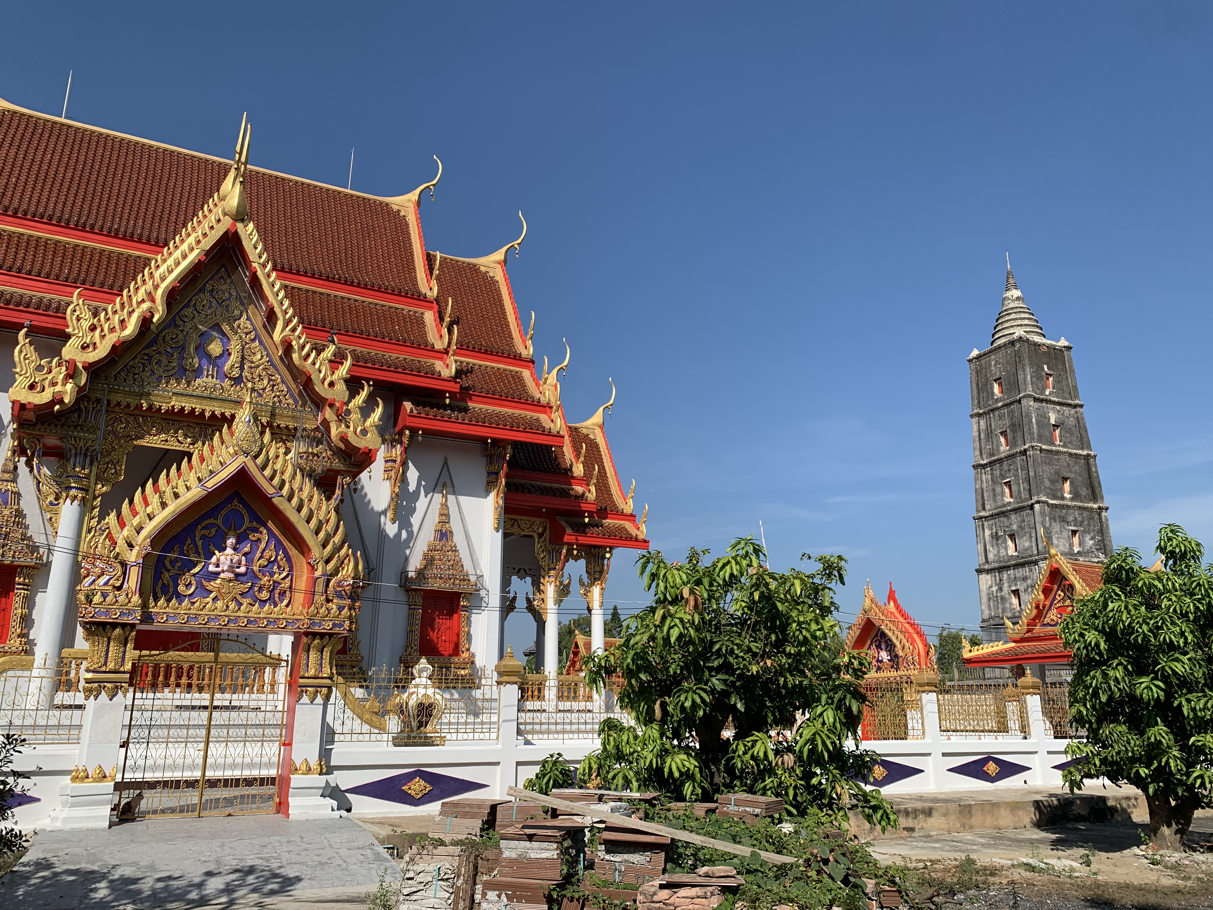 A sunlit view of Wat Tha It in Ang Thong, Thailand, featuring the ornate, white-walled temple with its red and gold roof on the left, contrasted against the tall, dark, multi-tiered chedi (stupa) on the right, all under a clear blue sky.