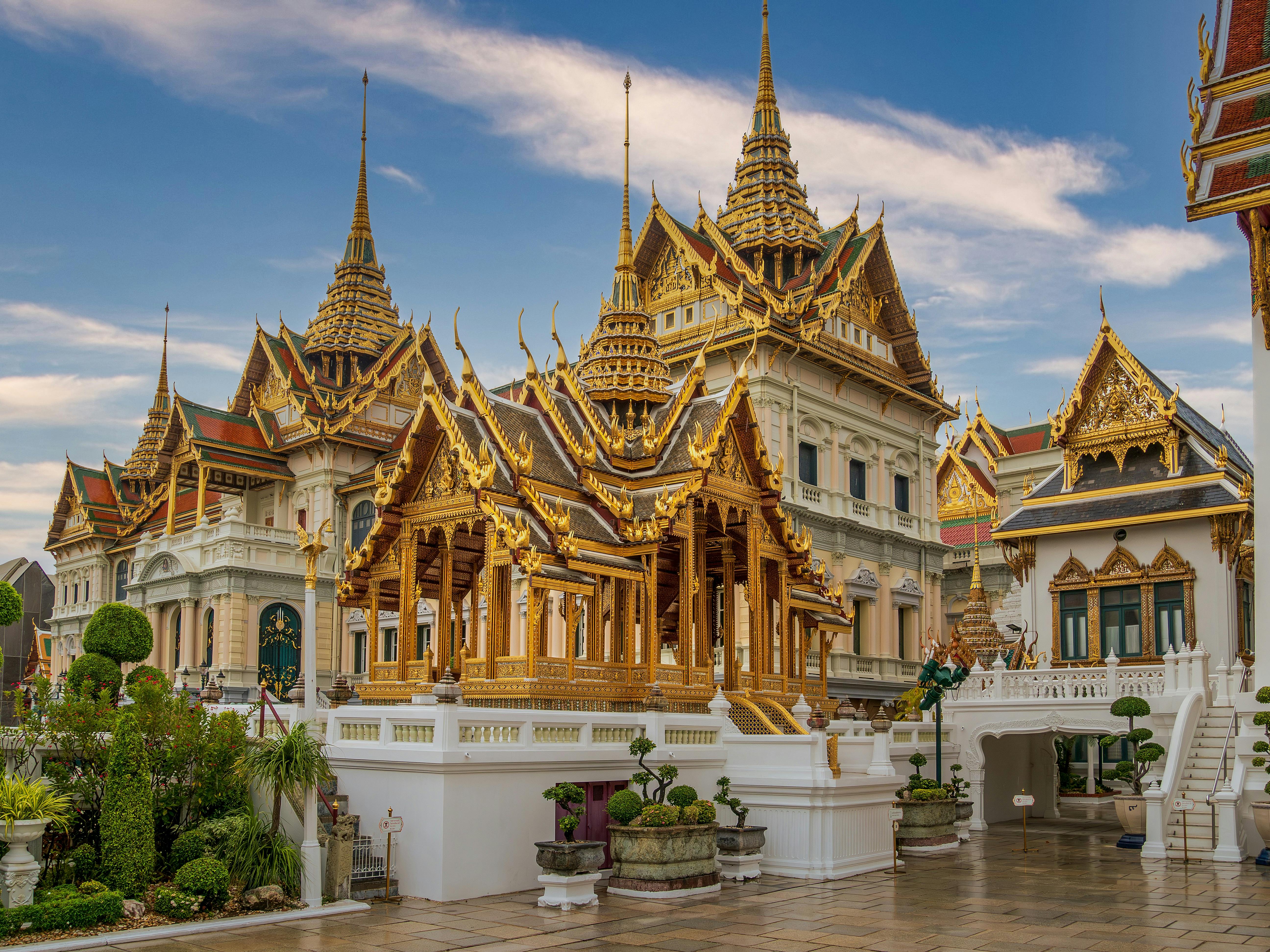An intricate, golden pavilion with a multi-tiered, spired roof stands in the courtyard of the Grand Palace complex in Bangkok, Thailand, with larger ornate halls and white walls in the background under a blue, cloudy sky.