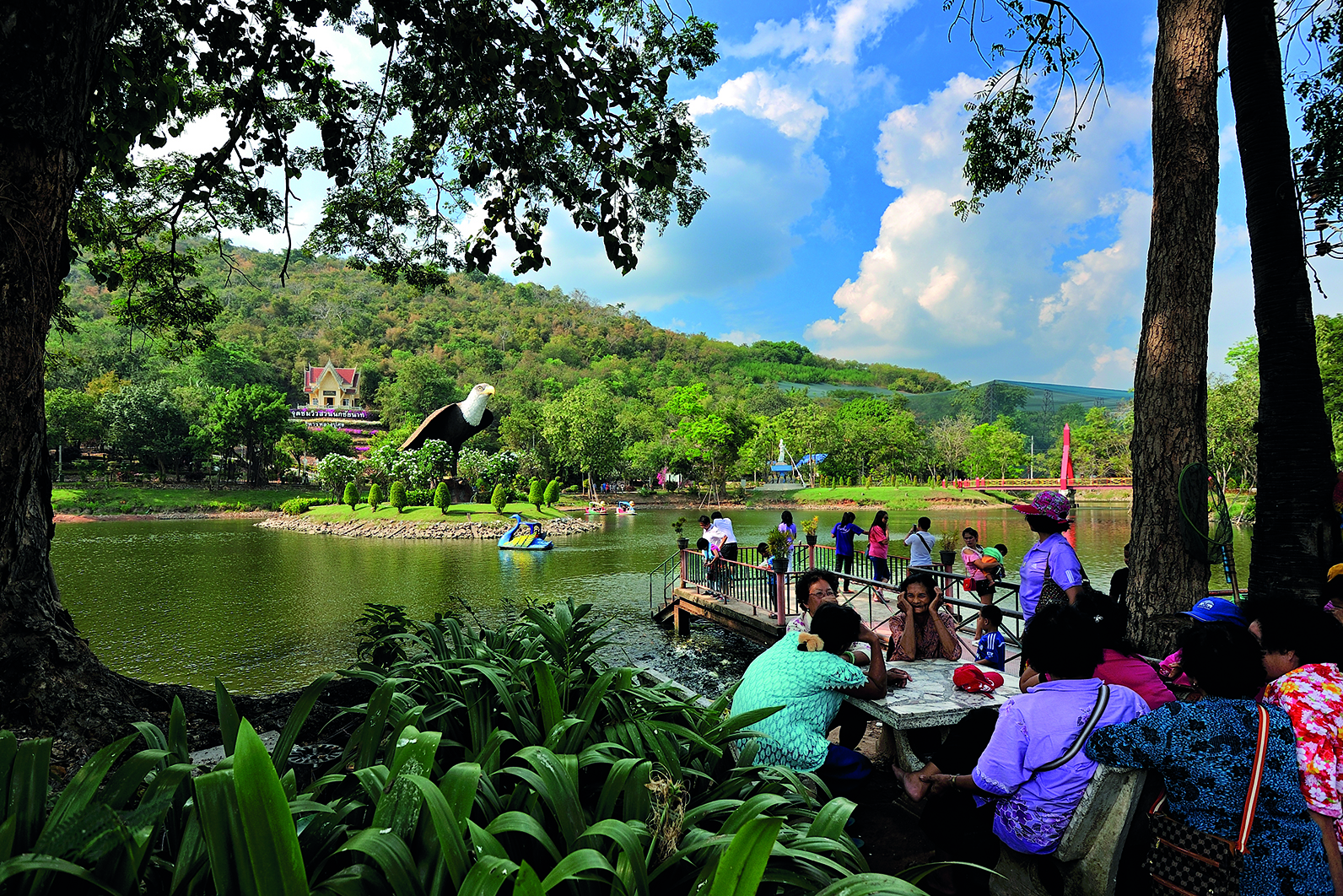 An outdoor daytime shot of the Chai Nat Bird Park in Thailand, showing visitors relaxing by a lake under a bright blue sky with fluffy clouds. In the foreground, a group of people sits at a white table on a wooden deck next to the water, shaded by a large tree on the left. Lush green plants frame the bottom of the image. In the middle ground, the lake stretches across the scene, with a massive, prominent statue of a bald eagle, wings partially spread, perched on a rock near the center. A small blue and yellow pedal boat is visible on the water. In the background, a forested green hillside rises up, with a multi-tiered, ornate Thai-style building with a red and gold roof visible among the trees on the left side of the hill.