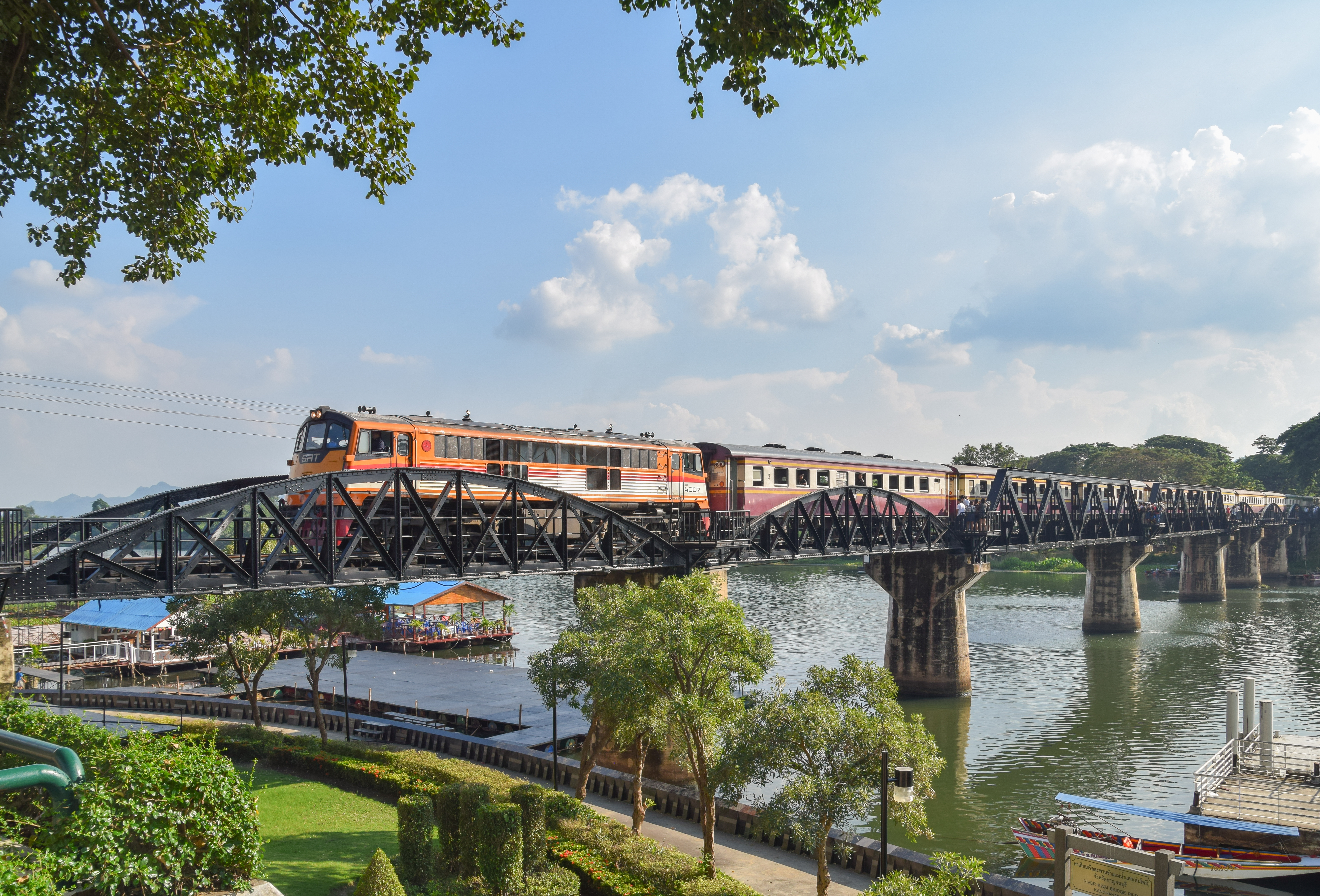 An orange and red train crosses the iconic Bridge on the River Kwai, a black metal trestle bridge, over the wide river in Kanchanaburi, Thailand, viewed from a lush, green bank under a blue sky with clouds.