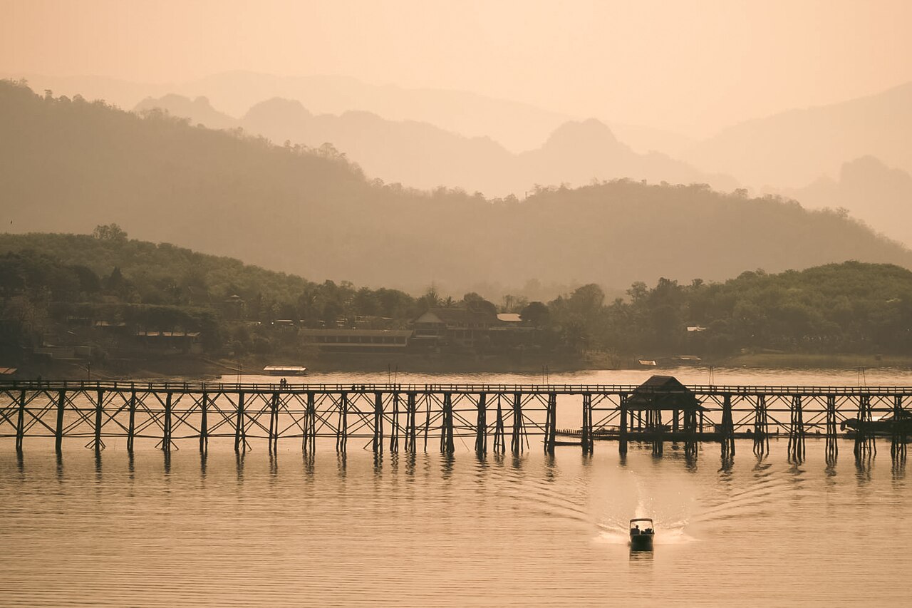 A serene, hazy view of the long, wooden Mon Bridge in Kanchanaburi, spanning a calm lake where a small boat is cruising. Layers of forest-covered mountains are visible in the background under a soft, warm light.