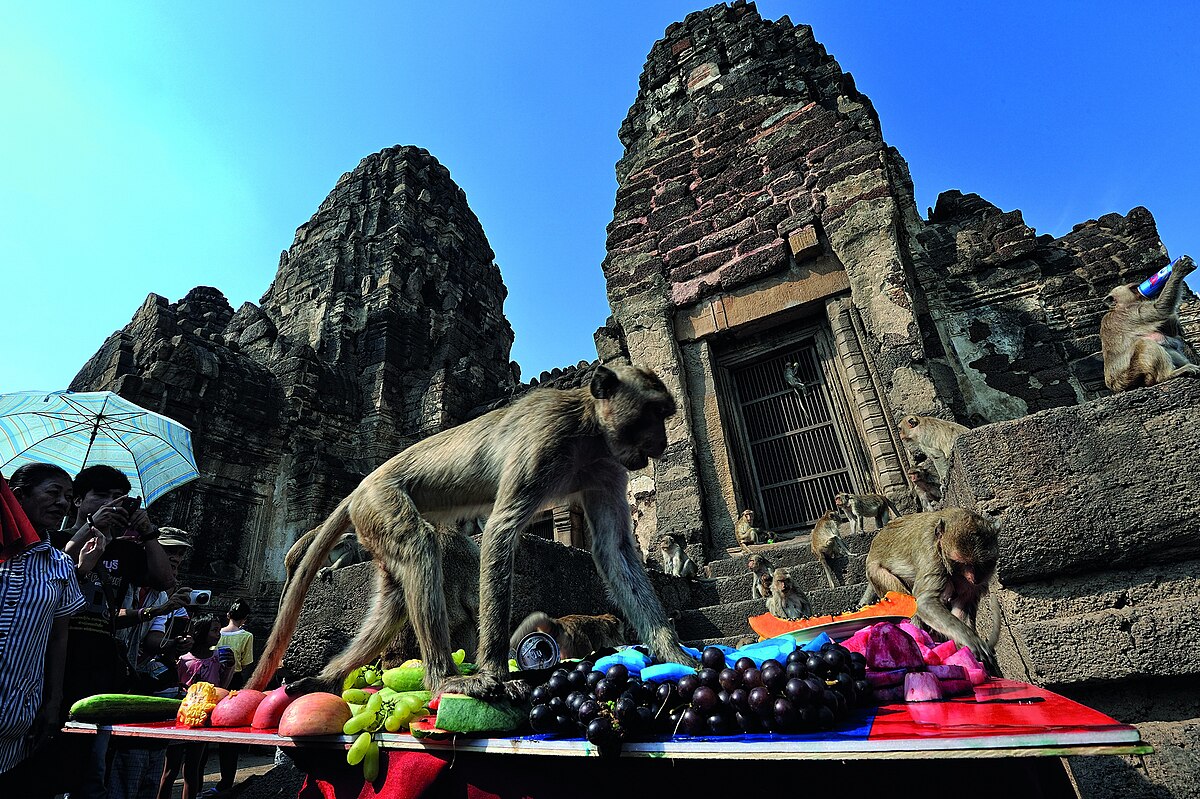 A low-angle, outdoor shot shows several macaques feasting on a table laden with fresh fruits, including grapes and watermelon, at the base of the ancient, towering stone ruins of Phra Prang Sam Yot temple in Lopburi, Thailand, under a clear blue sky.