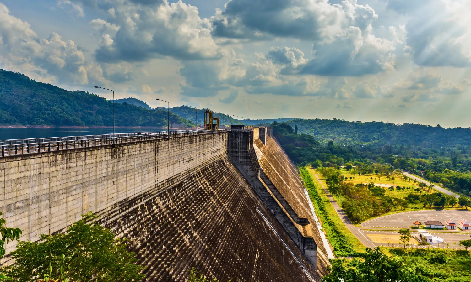 A high-angle, scenic view looking along the crest of the massive Khun Dan Prakan Chon Dam in Nakhon Nayok, Thailand, with its reservoir on one side and a lush green valley and forested hills on the other, under a partly cloudy sky.