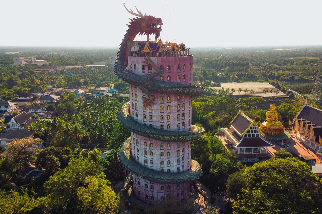 An aerial view of the Wat Samphran temple in Nakhon Pathom, Thailand, featuring a tall, cylindrical pink tower encircled by a massive, dark dragon sculpture that spirals from the base to the roof. To the right, a large, golden seated Buddha statue overlooks the surrounding lush, green landscape.