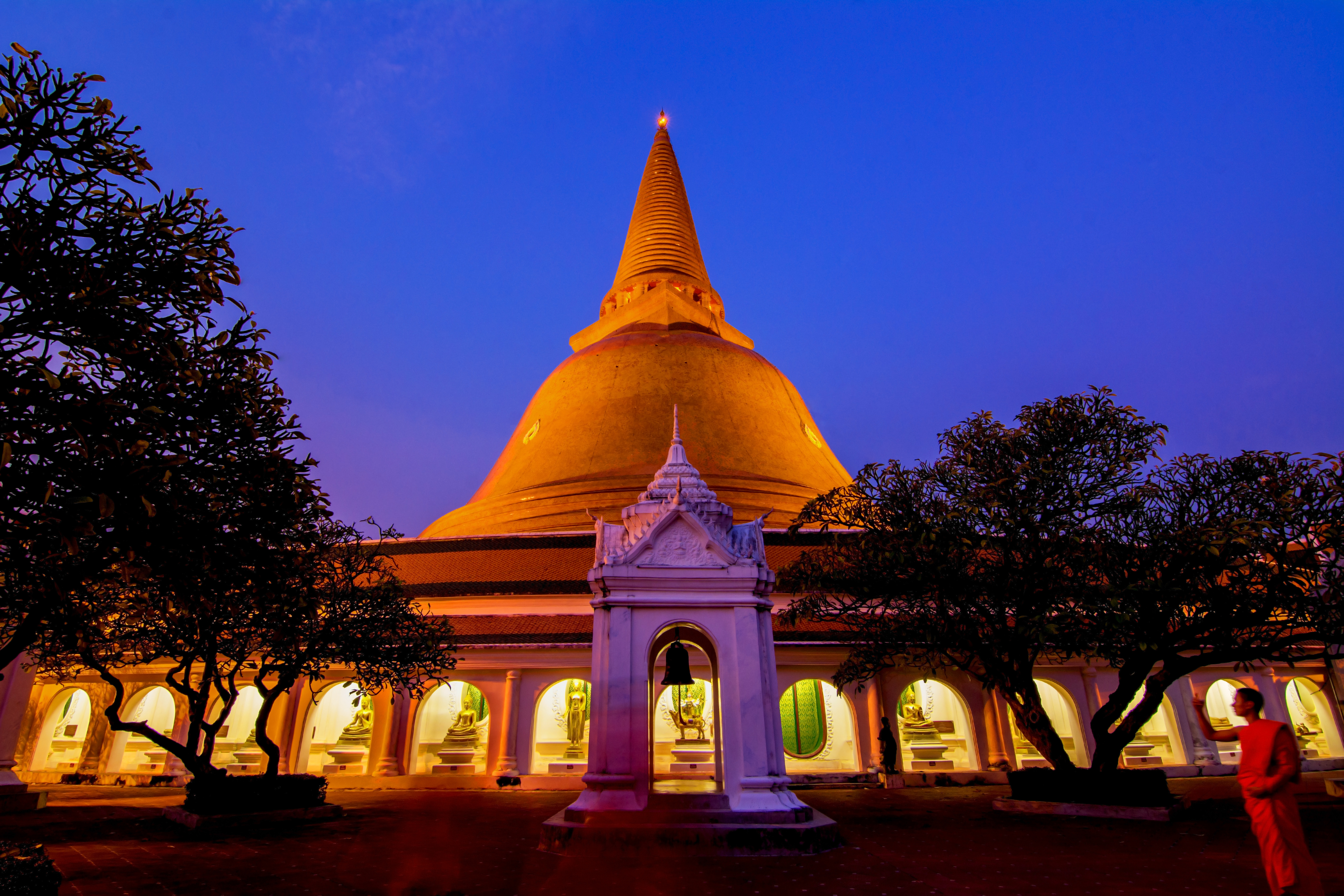 The magnificent golden stupa of Phra Pathom Chedi in Nakhon Pathom, Thailand, glows against a deep blue twilight sky, with a Buddhist monk in orange robes standing in the foreground courtyard.