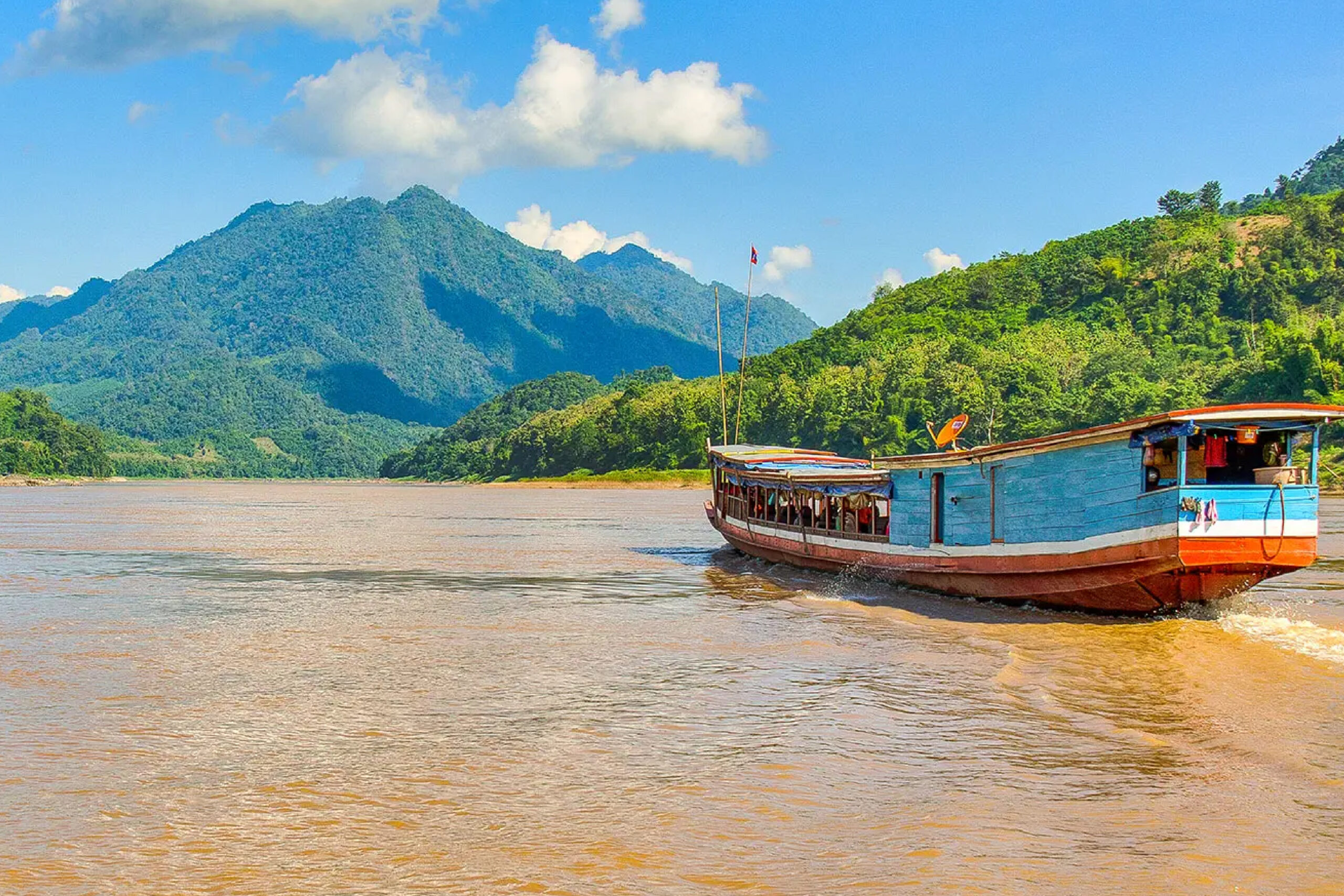 A thai cargo boat on muddy brown water surrounded by vibrant green jungle covered mountains.