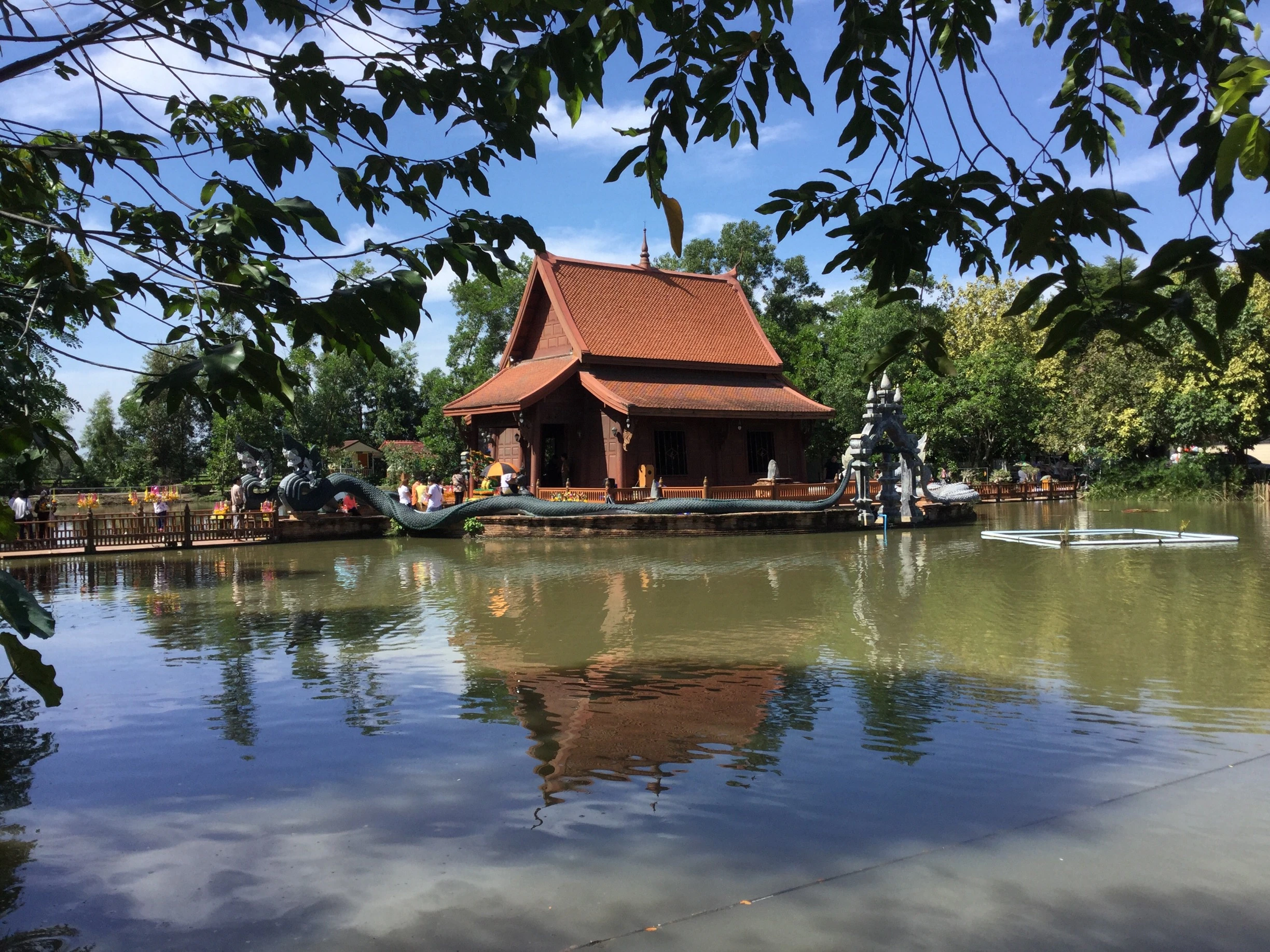A traditional Thai temple with an orange-tiled roof sits in the middle of a tranquil pond in Pathum Thani, Thailand. A large, ornate Naga serpent statue encircles the structure, its body forming a walkway leading to the entrance. The building and a blue sky are reflected in the water, and the scene is framed by dark green leaves in the foreground.
