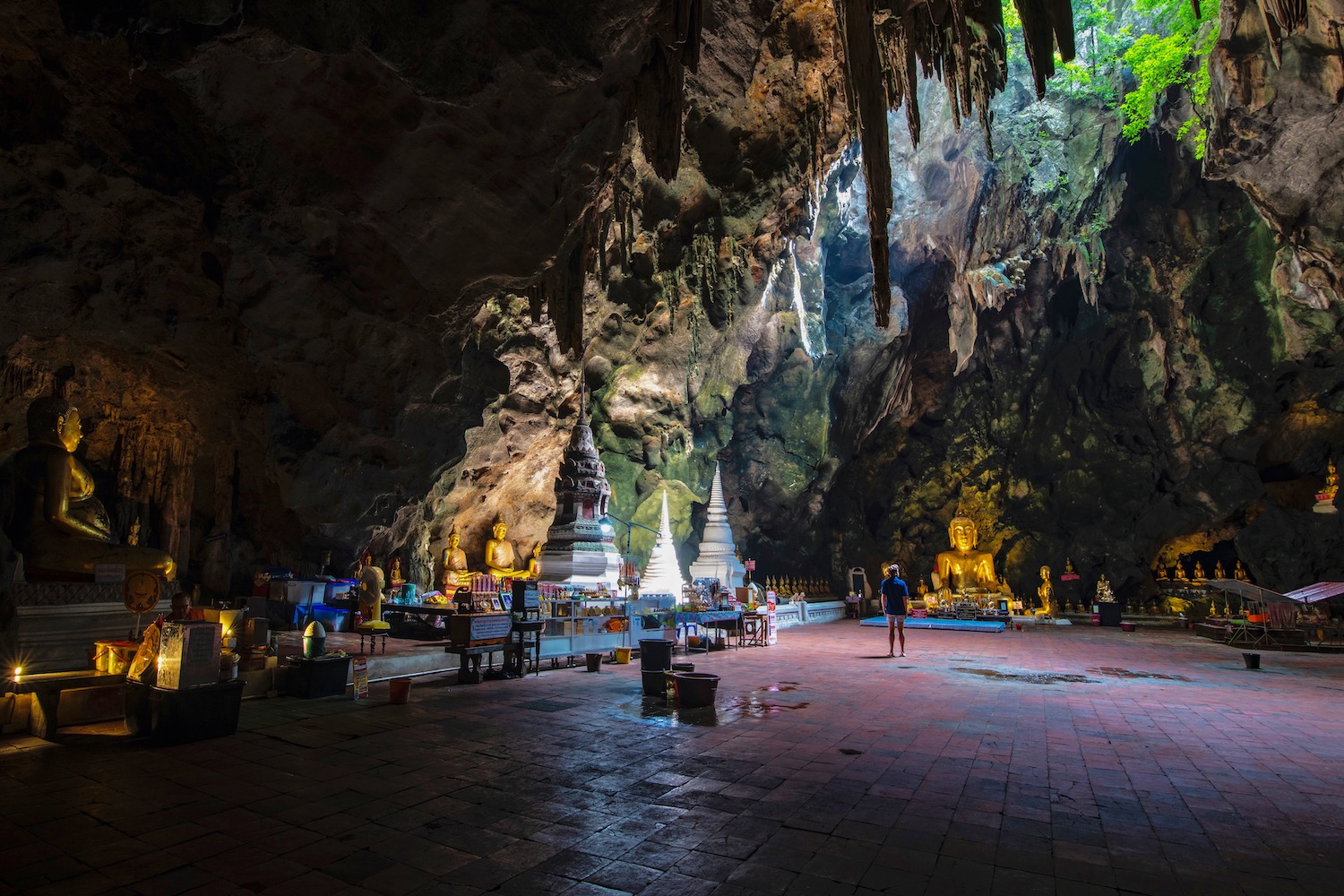 A wide-angle view inside the Tham Khao Luang cave temple in Phetchaburi, Thailand. The vast cavern is naturally lit by beams of sunlight shining down from an opening in the ceiling, illuminating several golden Buddha statues, two white stupas, and a person standing on the right.