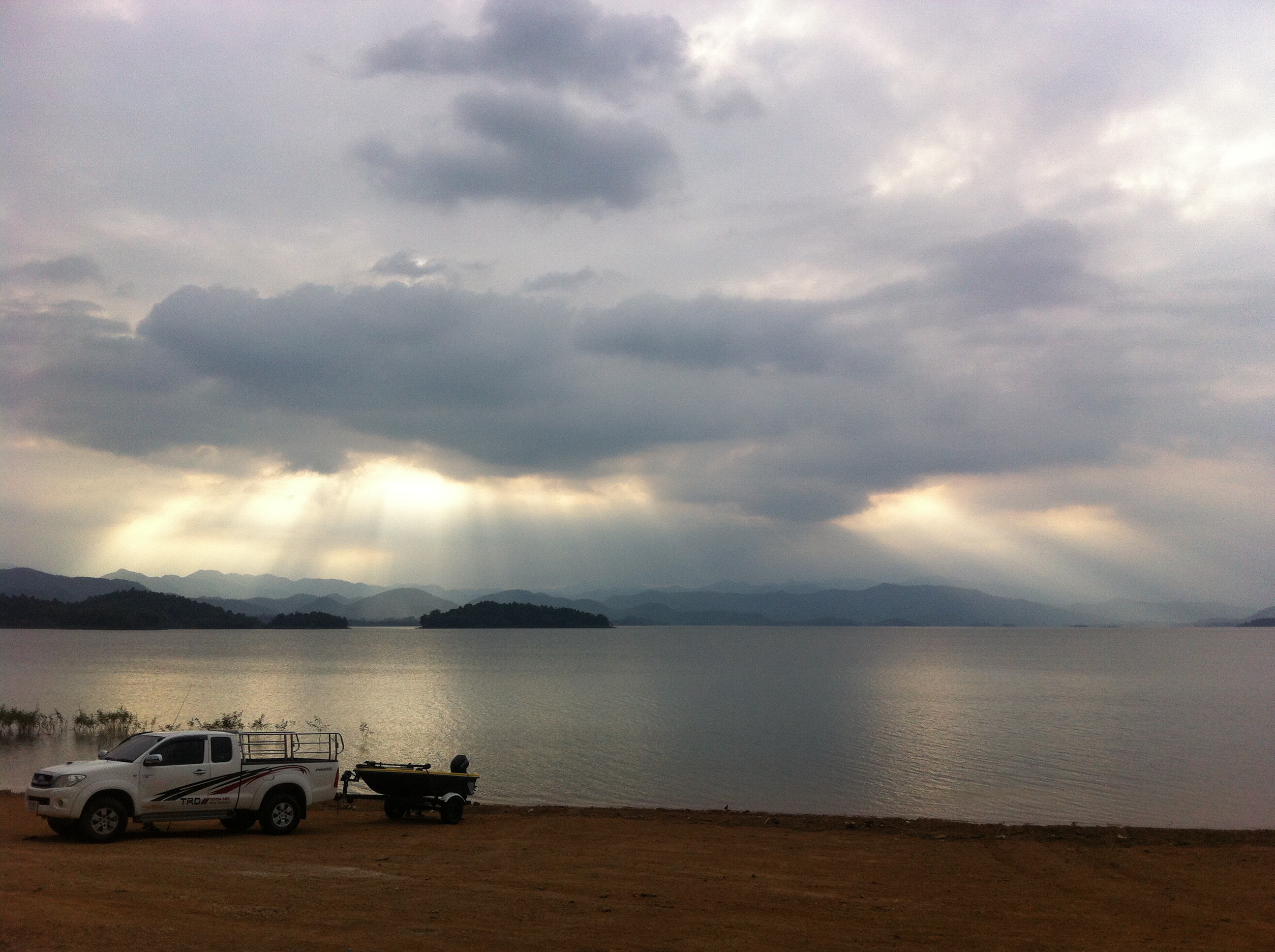 A white pickup truck with a boat trailer parked on the dirt shore of a large, calm lake in Phetchaburi, Thailand. In the distance, mountains line the horizon under an overcast sky, with dramatic sunbeams breaking through the clouds and shining onto the water.