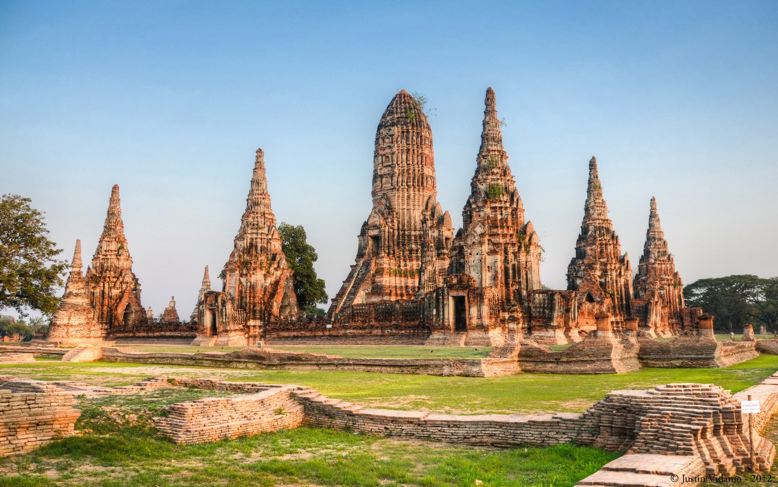 Landscape view of the ancient brick ruins of Wat Chaiwatthanaram in Phra Nakhon Si Ayutthaya, Thailand, featuring a central Khmer-style prang tower surrounded by smaller spires under a clear blue sky.