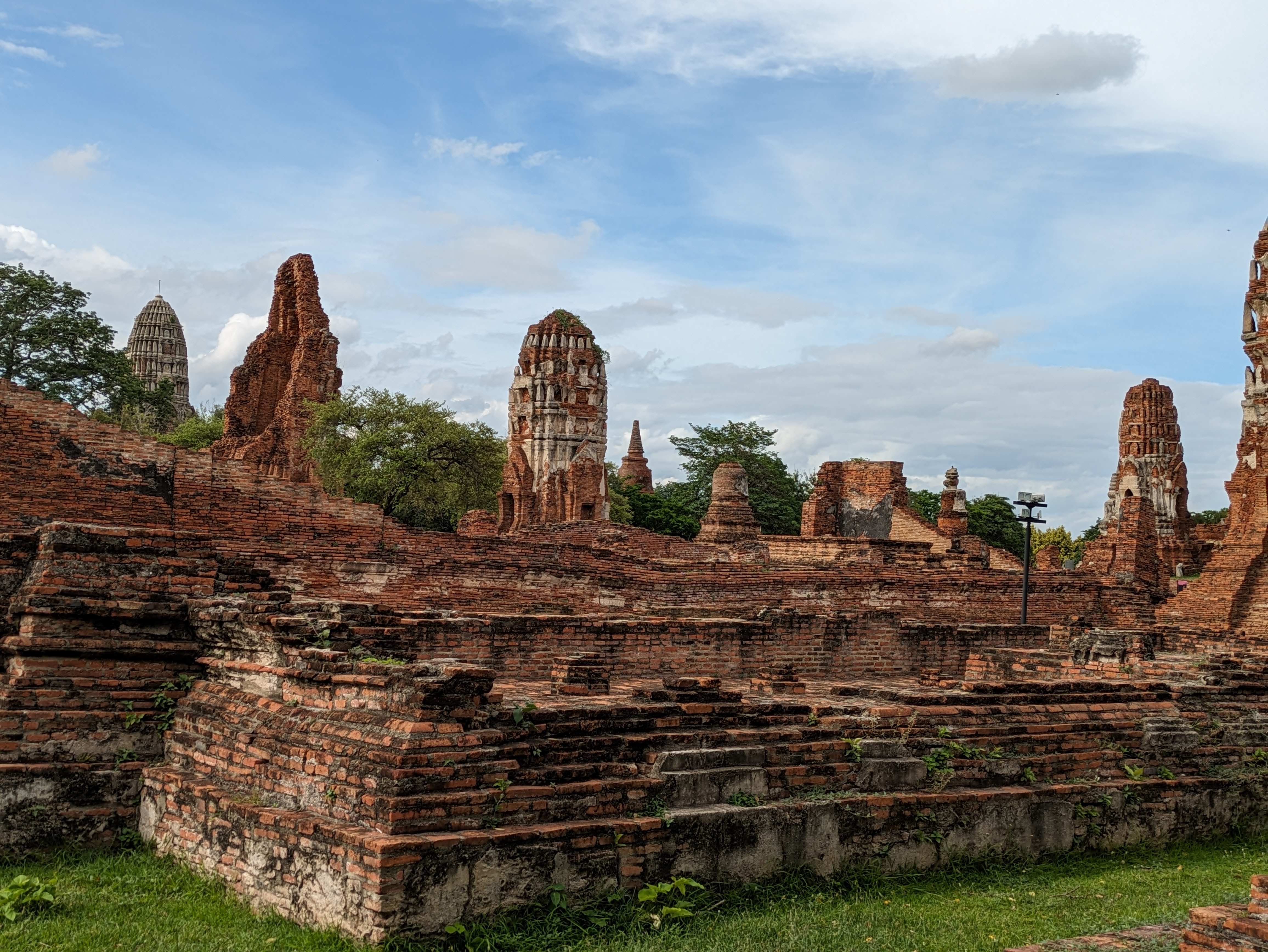 The red brick ruins of the ancient temples at Ayutthaya Historical Park in Thailand, with crumbling walls in the foreground and several towering prangs against a blue sky with clouds.