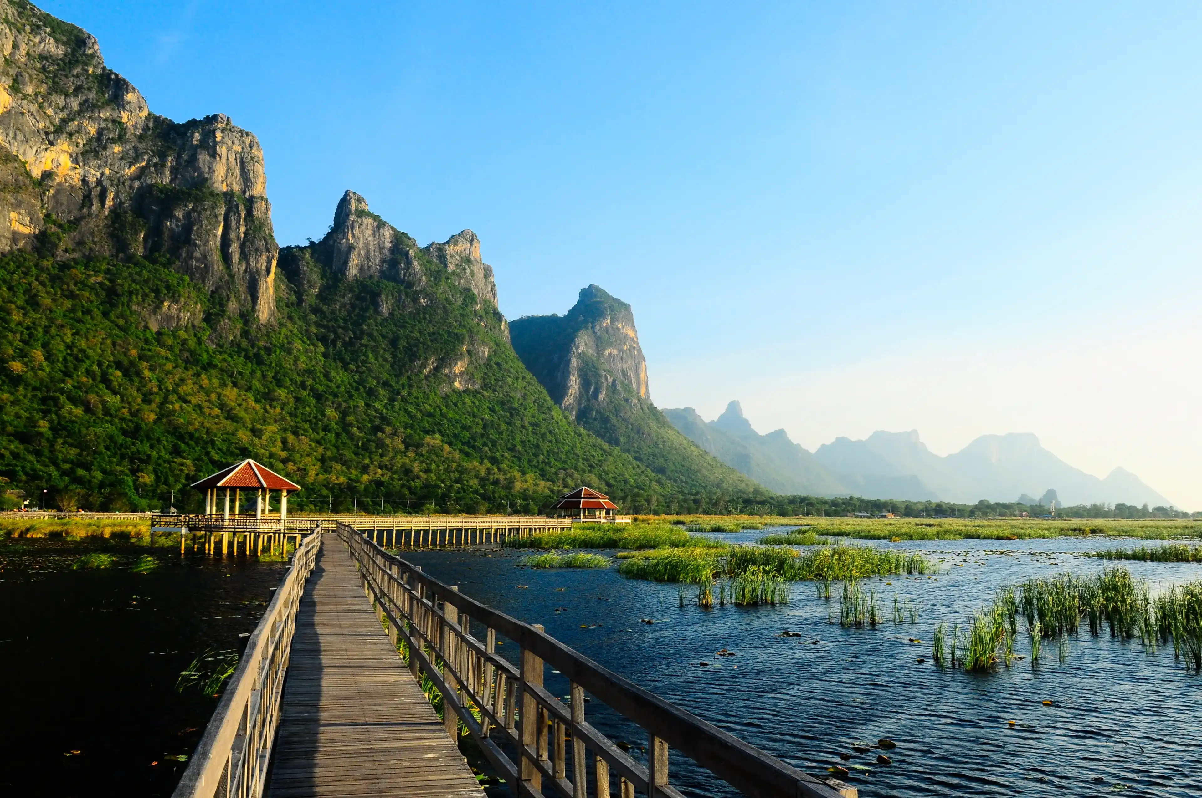 A scenic wooden boardwalk stretching across a freshwater marsh filled with green reeds, leading toward covered pavilions with dramatic limestone mountains rising in the background at Khao Sam Roi Yot National Park in Prachuap Khiri Khan, Thailand.