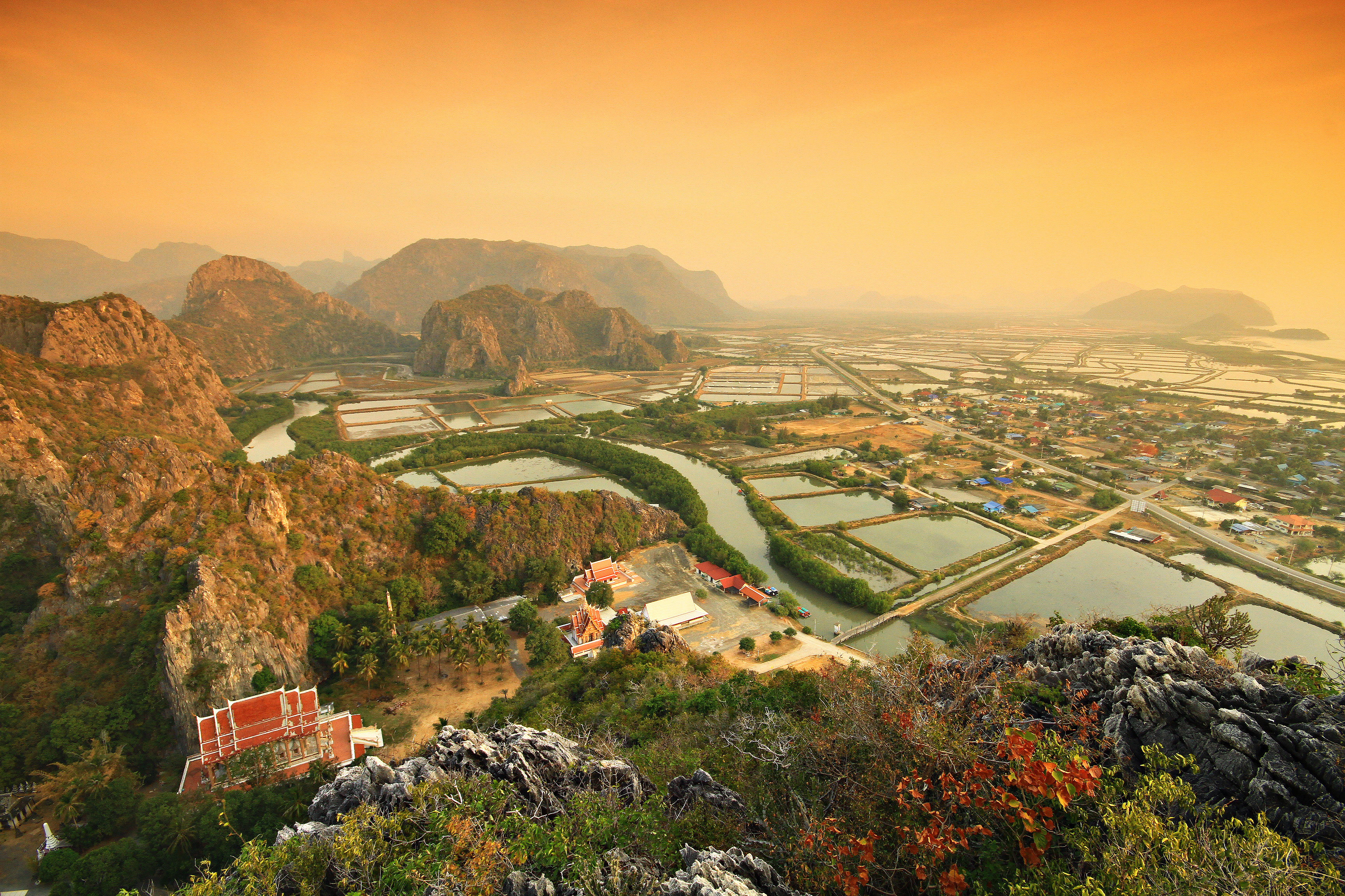 An elevated, panoramic view of Prachuap Khiri Khan province in Thailand at sunset. A red-roofed temple is nestled in the foreground, overlooking a vast landscape of winding rivers, aquaculture farms, and a small town, with dramatic limestone mountains rising in the hazy, golden-lit background.