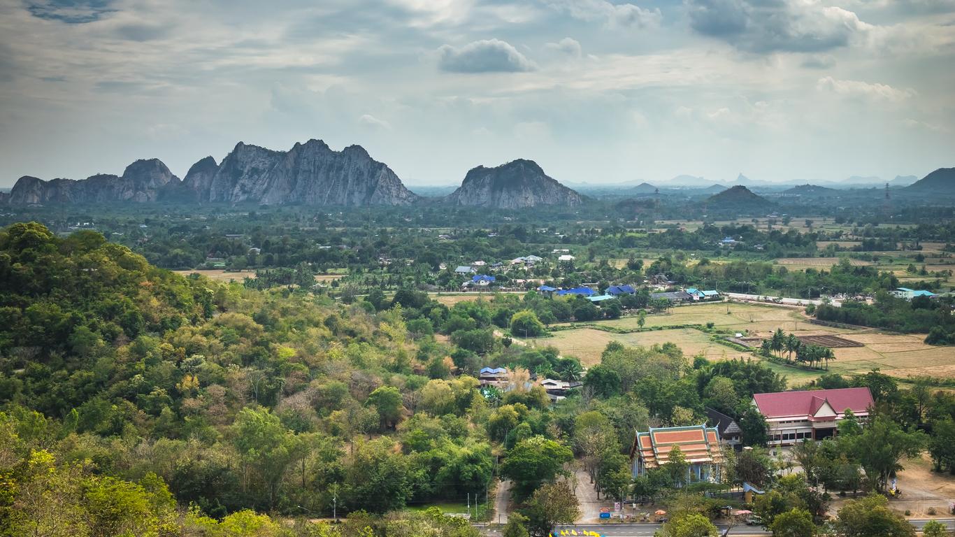 Panoramic view of the rural Ratchaburi landscape featuring dramatic limestone karst mountains rising above green forests, fields, and a village with a traditional red-roofed Thai temple.
