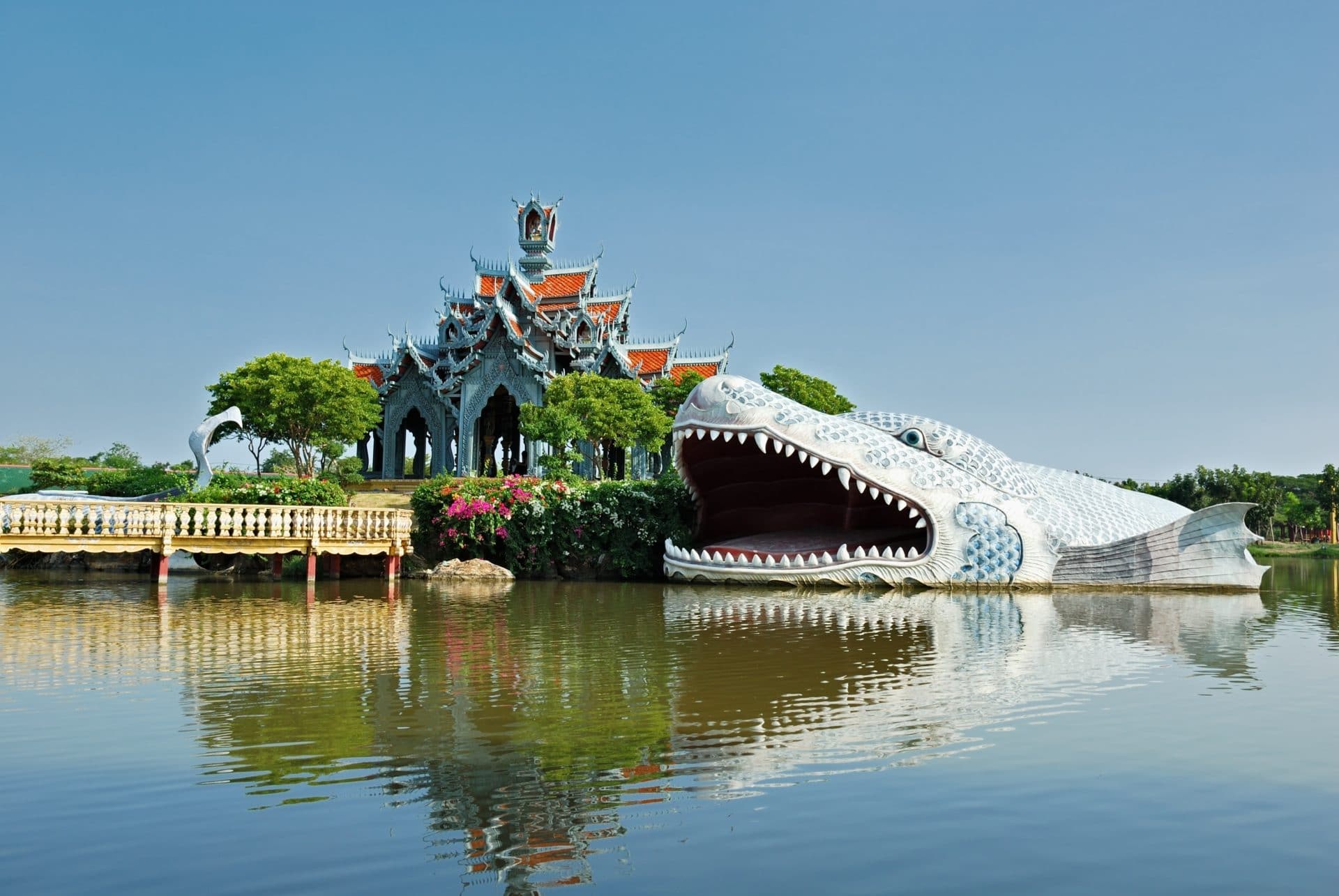 A giant mythical fish sculpture with an open mouth emerging from the water in front of an ornate Thai pavilion at the Ancient City (Muang Boran) in Samut Prakan, Thailand.
