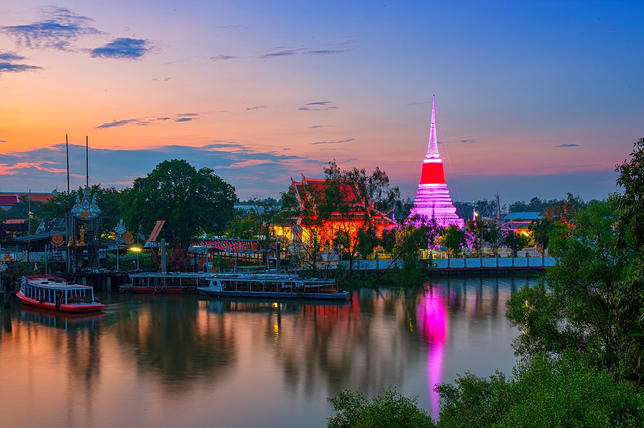 A riverside view of Phra Samut Chedi in Samut Prakan, Thailand, at sunset. The chedi is brightly illuminated in pink and white, reflecting on the calm water next to a traditional temple and docked boats under a colourful sky.