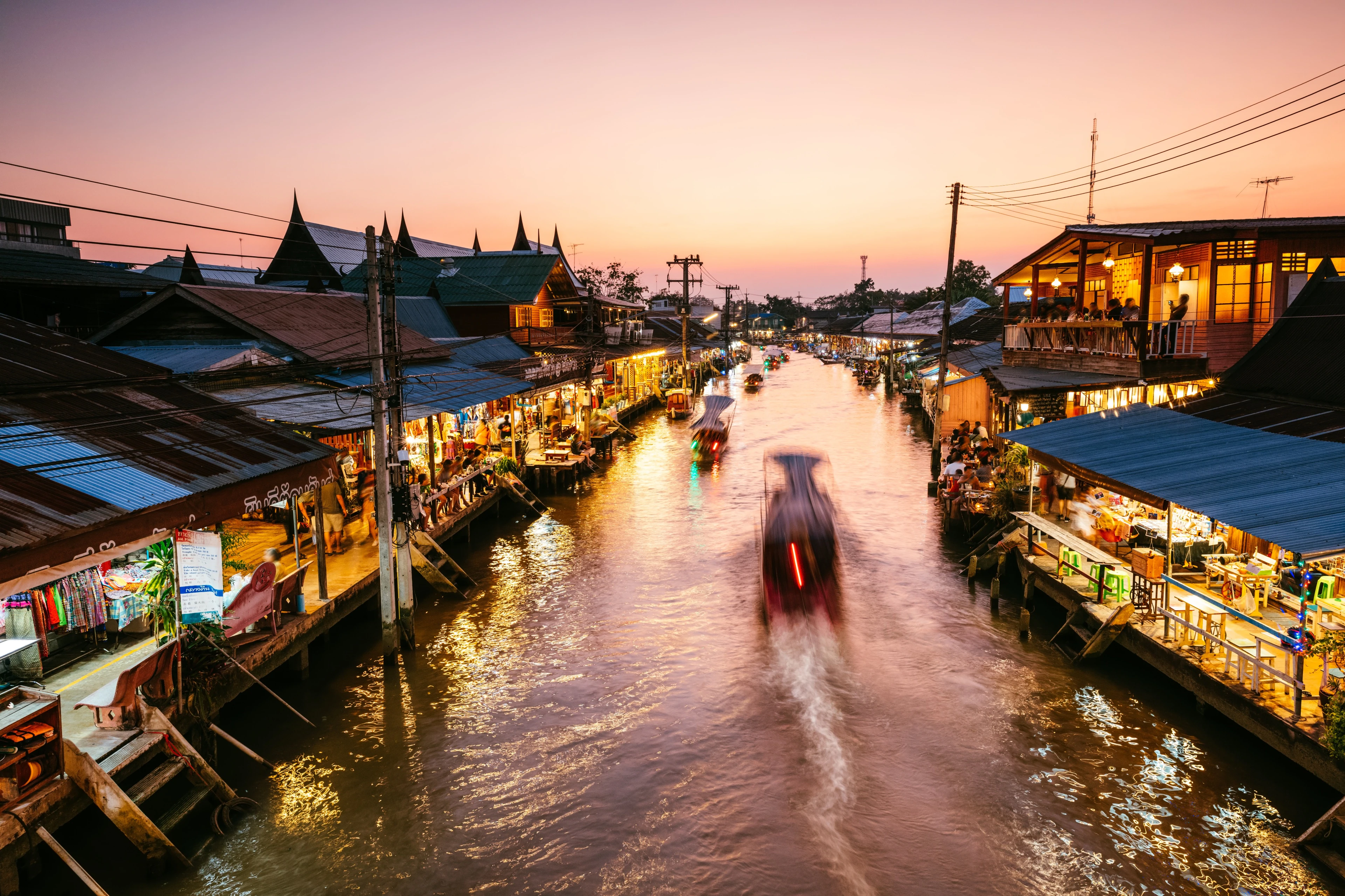 Scenic twilight view of a floating market canal in Samut Sakhon, Thailand, featuring illuminated wooden stilt houses lining the water and motion-blurred boats navigating the river.