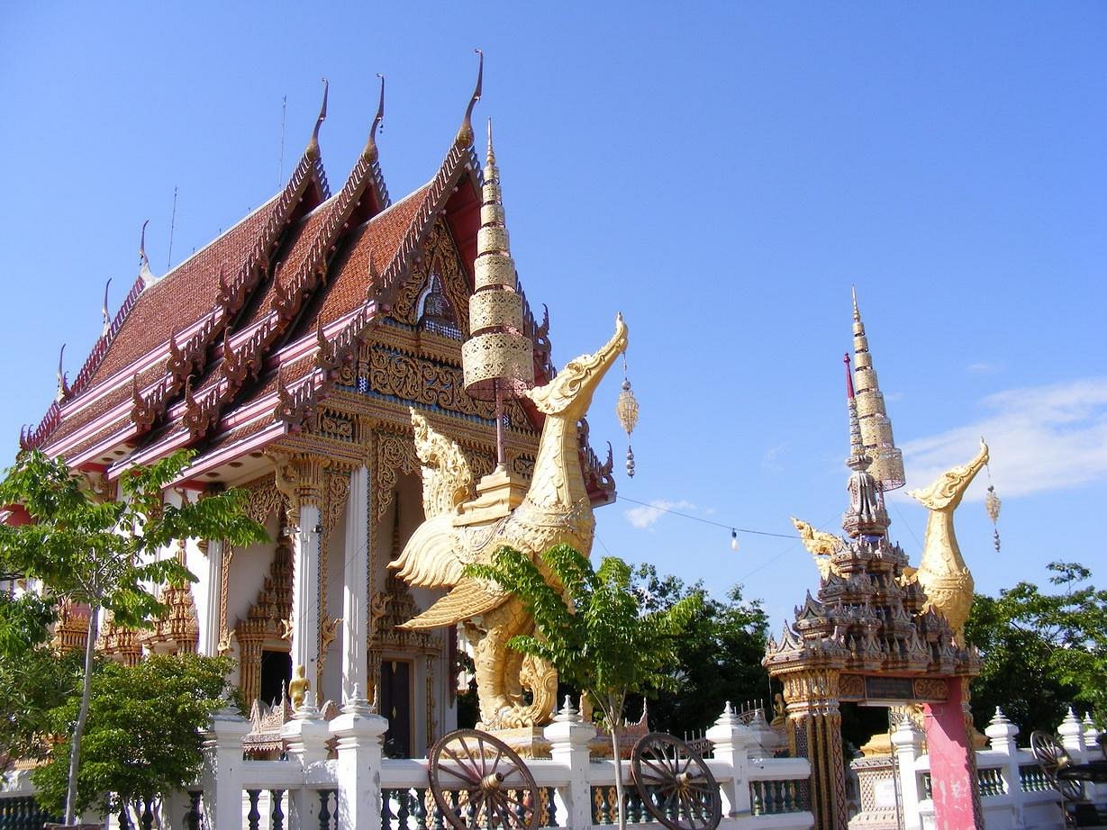 A large, golden statue of a mythical bird stands before an ornate Thai temple in Samut Sakhon, featuring a multi-tiered red and gold roof, all set against a clear blue sky.
