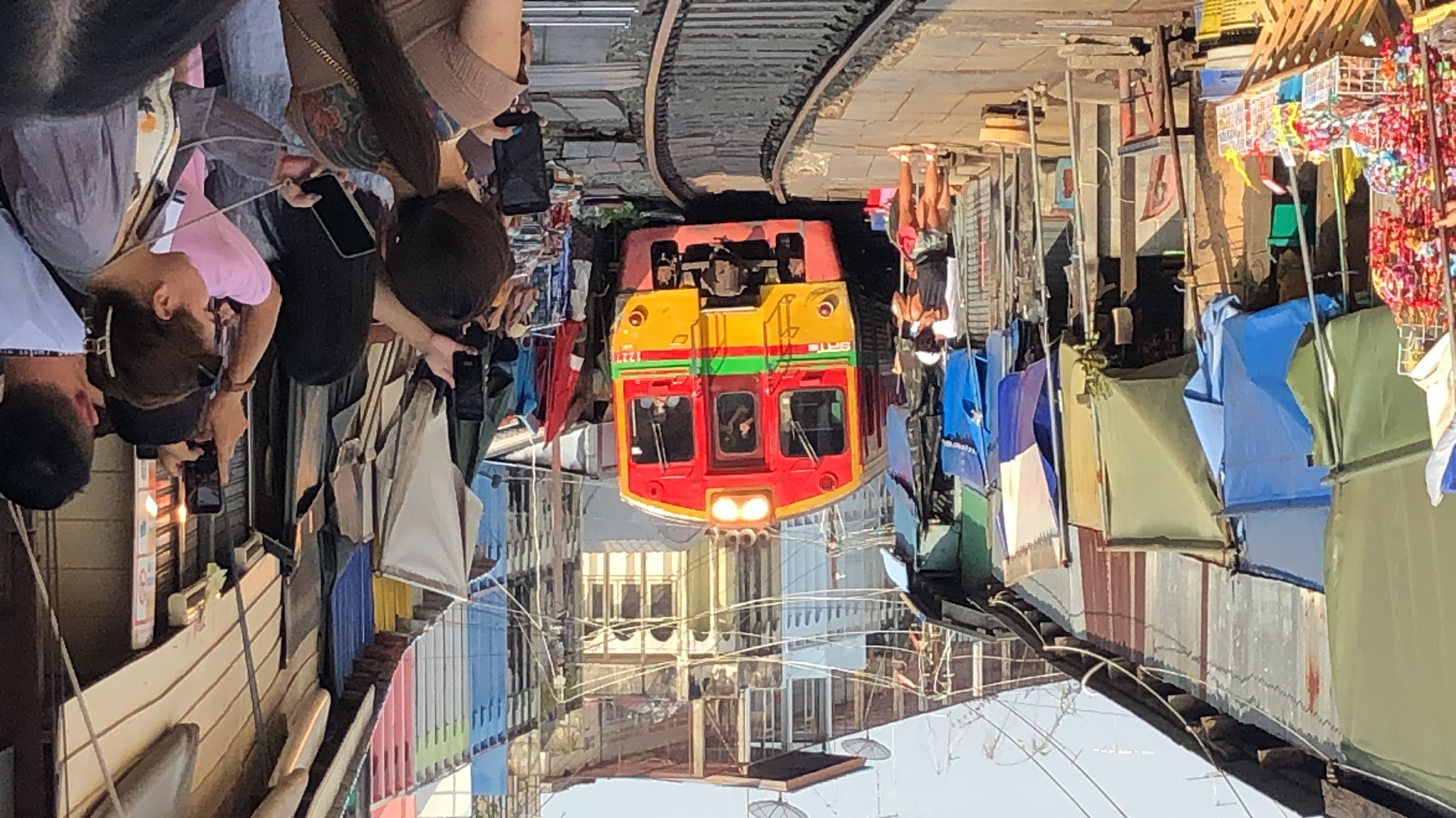 A red and yellow train approaches on a track running directly through the center of a bustling market in Samut Songkhram, Thailand. Crowds of tourists stand inches from the track, holding up their phones to photograph the train as it passes the colourful market stalls.