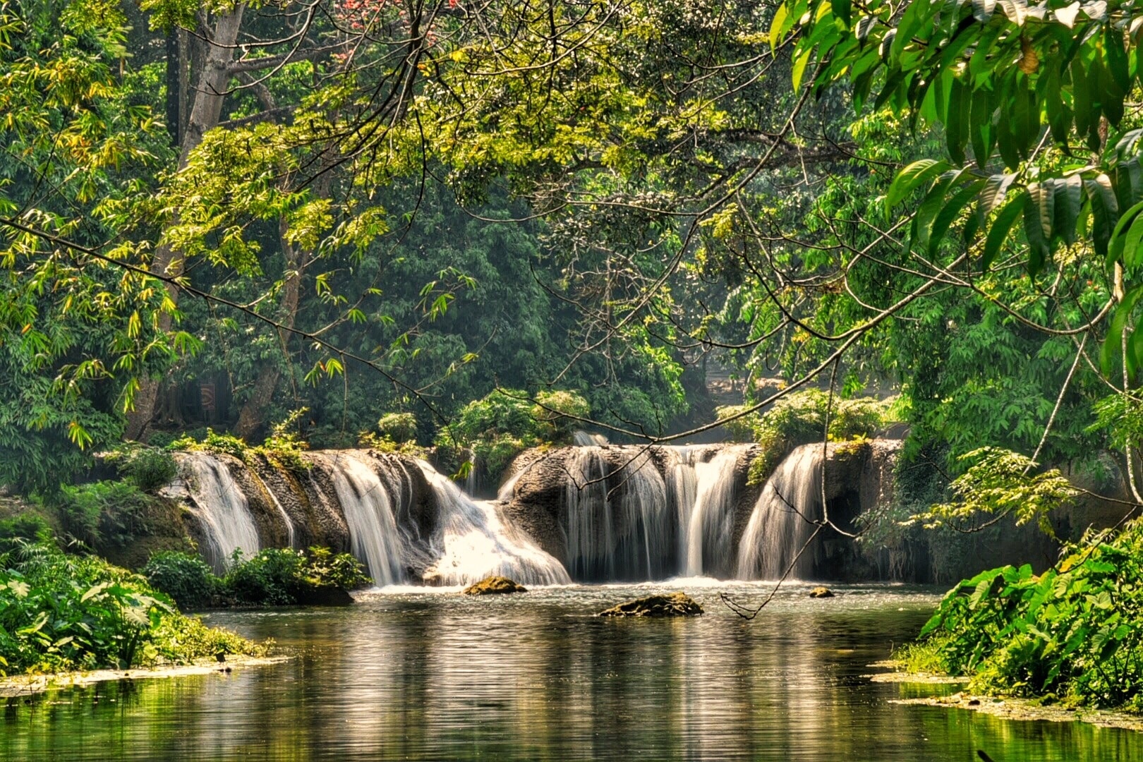 Scenic waterfall in Saraburi province, Thailand, featuring multiple cascades flowing over smooth rocks into a calm, reflective stream, framed by lush tropical foliage and sunlit overhanging branches.