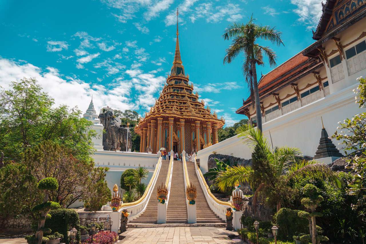 A wide stone staircase with ornate Naga serpent railings leads up to the magnificent golden Mondop of Wat Phra Phutthabat temple in Saraburi, Thailand, under a bright blue sky with white clouds.