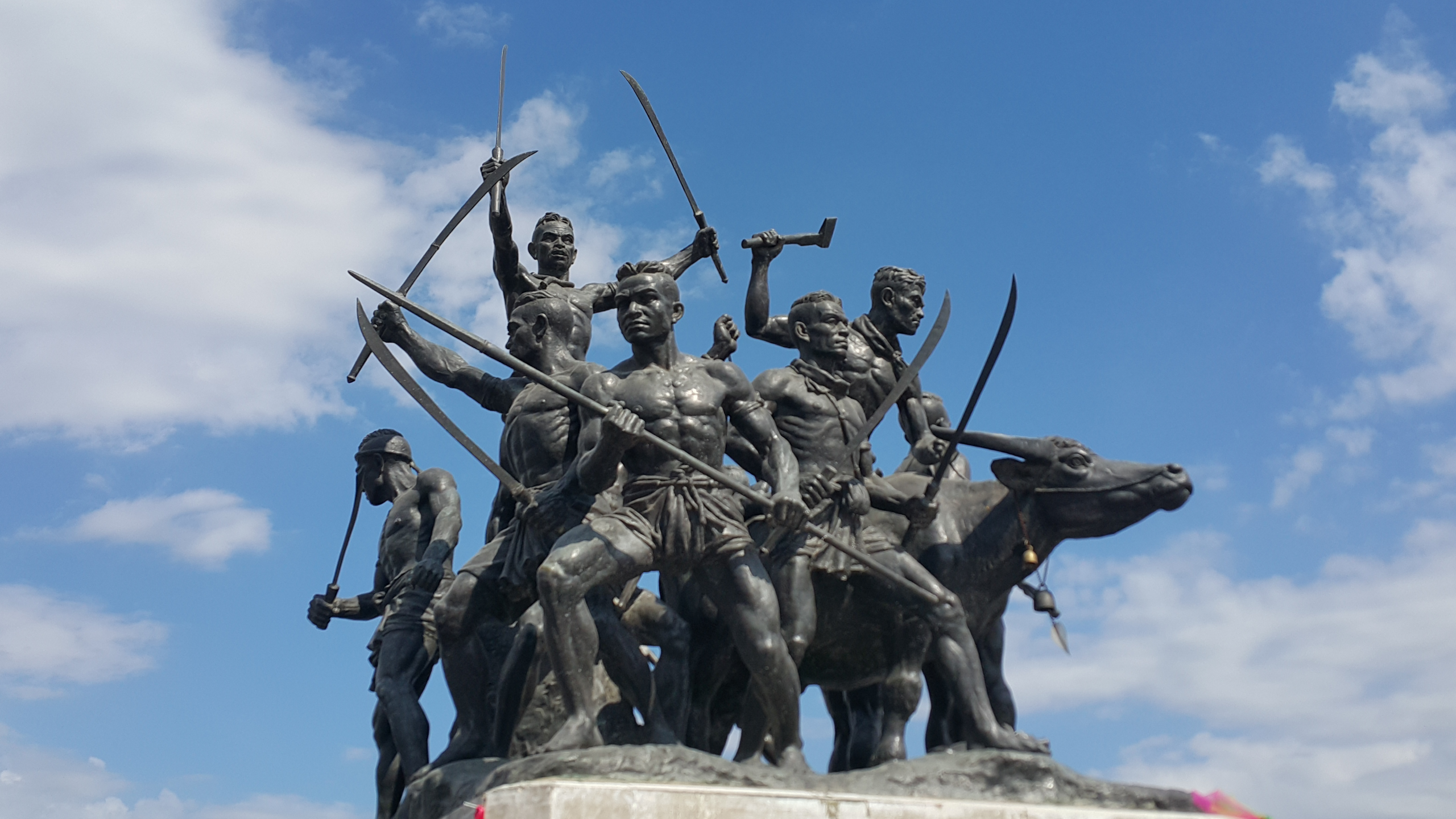 A low-angle photograph captures the bronze Monument of Bang Rachan Heroes in Sing Buri, Thailand, set against a bright blue sky with scattered white clouds. The dynamic sculpture depicts a group of shirtless warriors in traditional attire, charging forward with swords, spears, and axes raised. A large water buffalo stands alongside the men, also appearing to join the charge, adding to the scene's sense of movement and determination.
