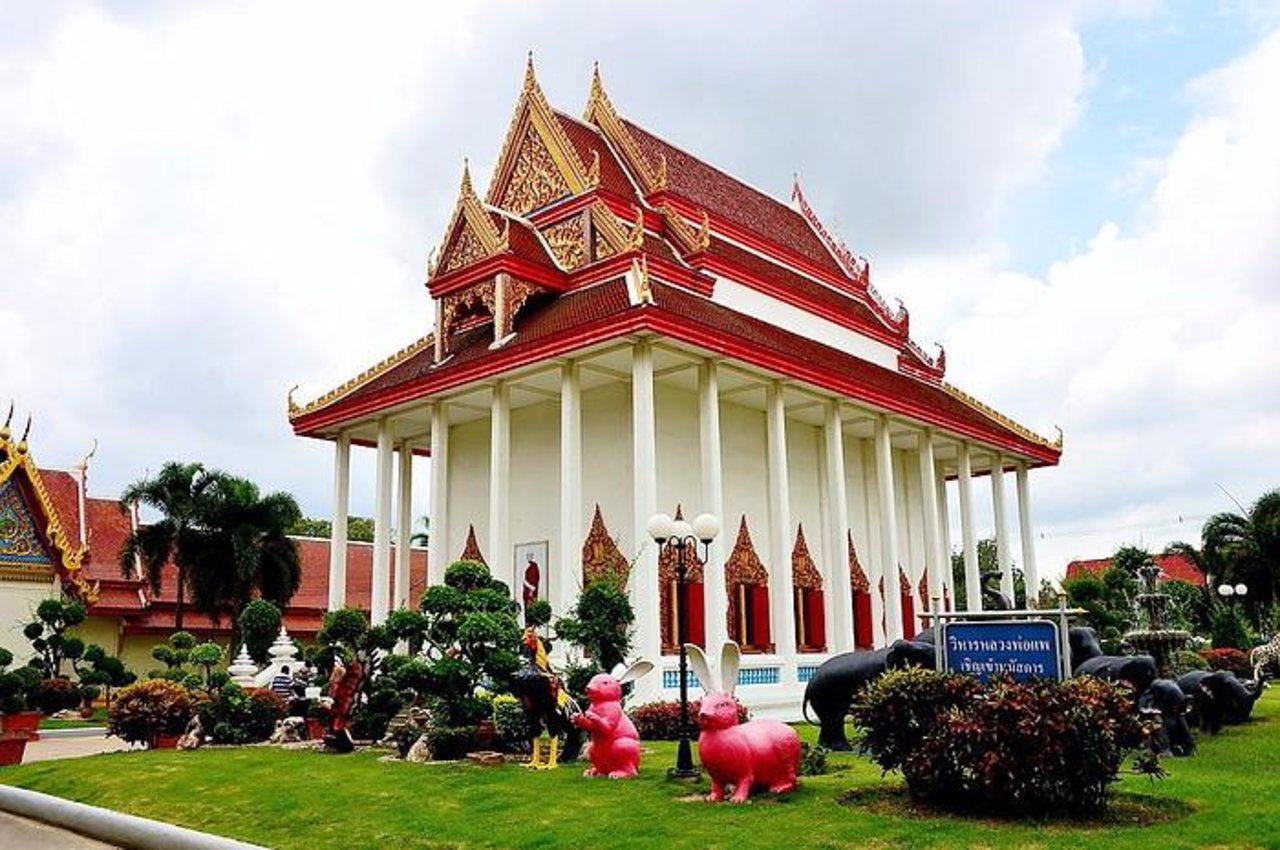 A white Thai temple in Sing Buri with an ornate, multi-tiered red and gold roof. In the foreground, on a manicured green lawn, are several colourful and whimsical statues, including two large pink rabbits, a rooster, and a black elephant.