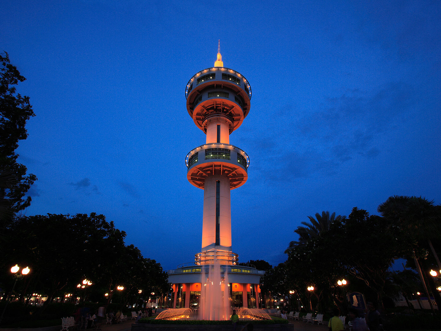 A low-angle, night view of the illuminated Banharn-Jamsai Tower in Suphan Buri, Thailand. The tall, white observation tower features multiple circular decks and is lit with warm orange lights against a deep blue twilight sky. At the base, a fountain sprays water, surrounded by a park area with dark trees, streetlamps, and benches.