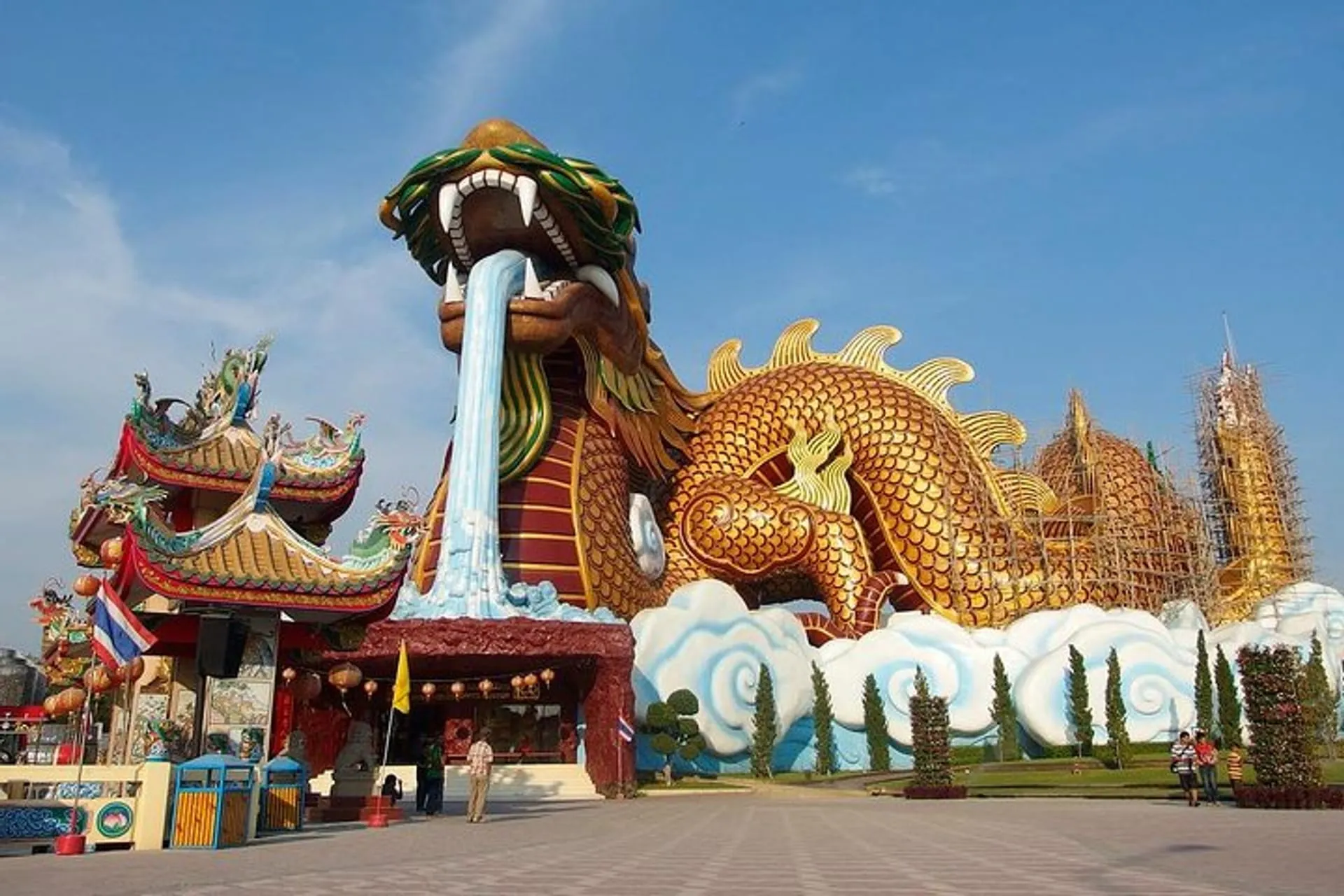 A massive, golden dragon statue at the Dragon Descendants Museum in Suphan Buri, Thailand. The dragon is coiled over stylized clouds with its mouth wide open, spouting a cascade of water onto a structure below. Visitors walk on the paved plaza in front of the immense statue under a clear blue sky.